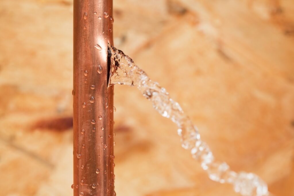 A household 1/2" copper water pipe has frozen and cracked. Water is leaking from the crack into an unfinished basement area, the background is oriented strand board (OSB).